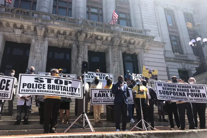 Newark Mayor Ras Baraka on the steps of City Hall surrounded by banners that say "We want justice" and "stop police brutality"